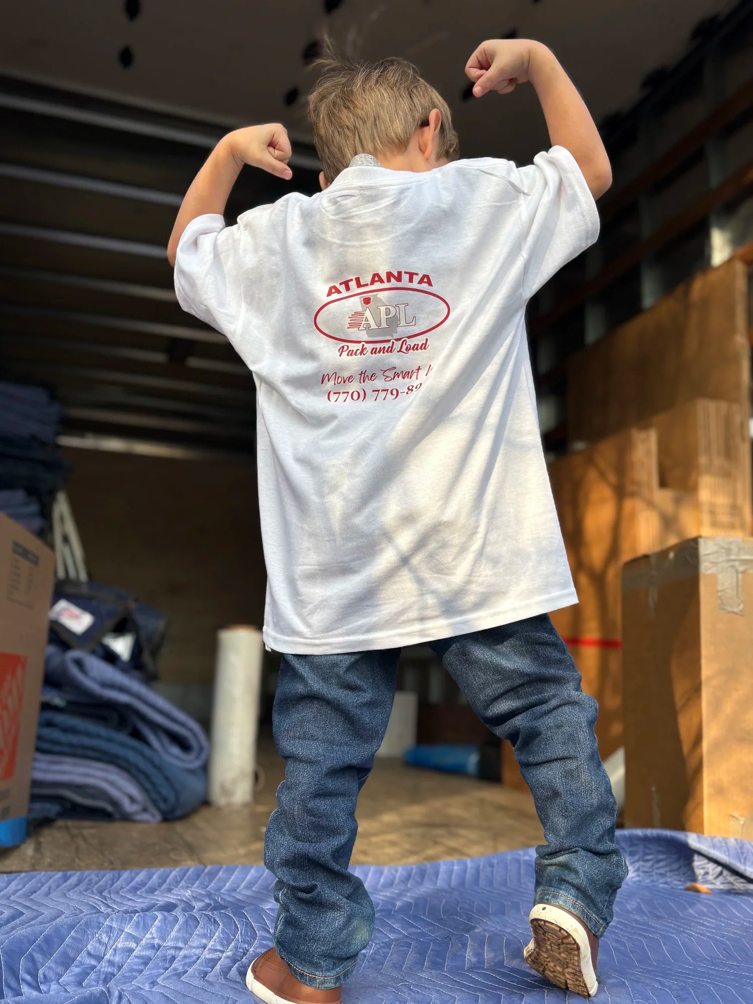 Child in Atlanta Pack and Load branded shirt flexing muscles inside moving truck, surrounded by packing blankets and boxes, symbolizing a stress-free moving experience.