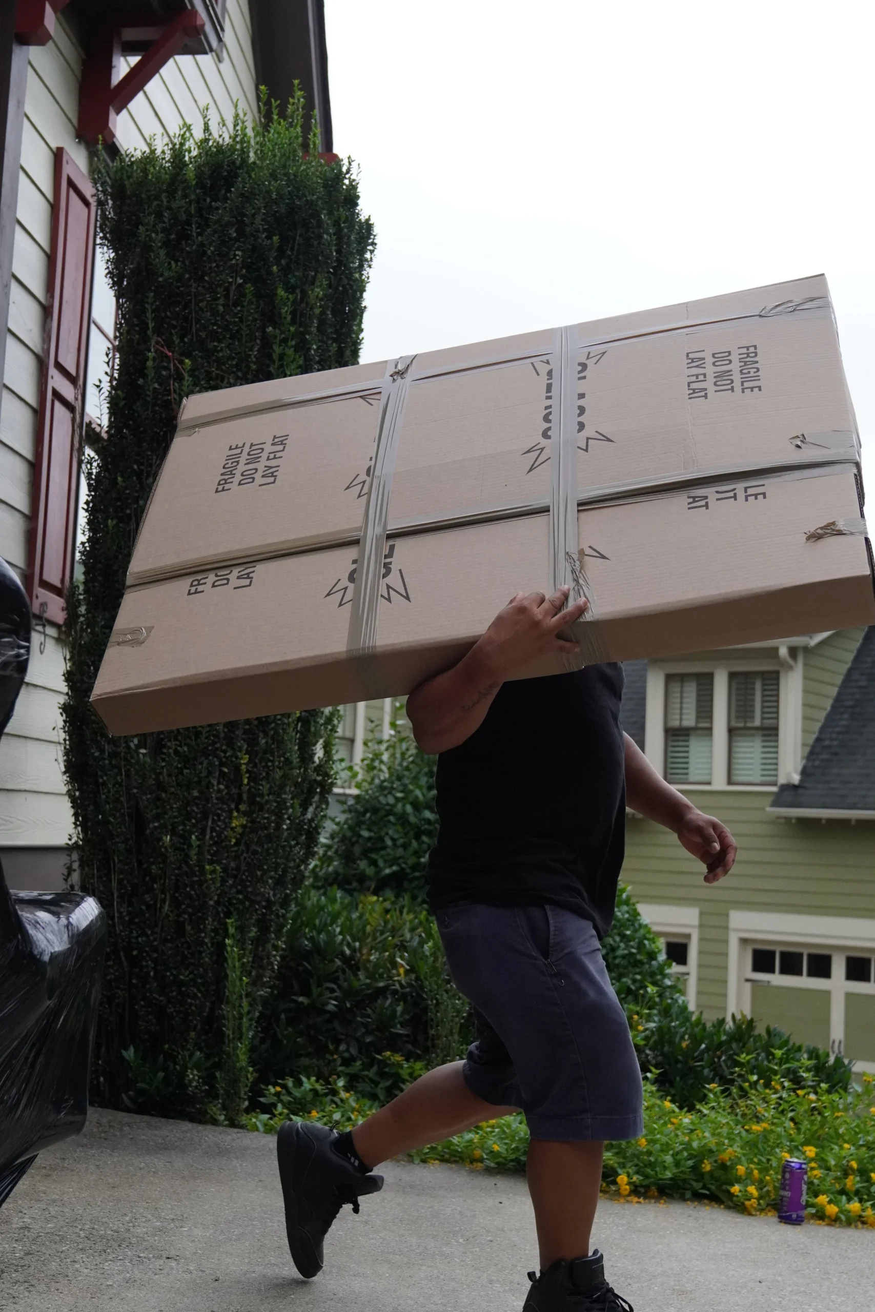 Person carrying a large, fragile box labeled "Do Not Lay Flat" while walking on a driveway surrounded by greenery, illustrating the physical demands of local moving services in Austell, GA.