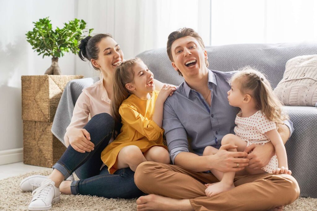 Family enjoying quality time together in a cozy living room, smiling and playing on the floor, surrounded by a comfortable couch and indoor plants.