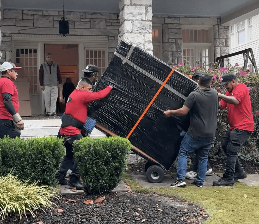 Four movers in red shirts transporting a large, wrapped item on a dolly outside a house, demonstrating Atlanta Pack and Load's long-distance moving services.