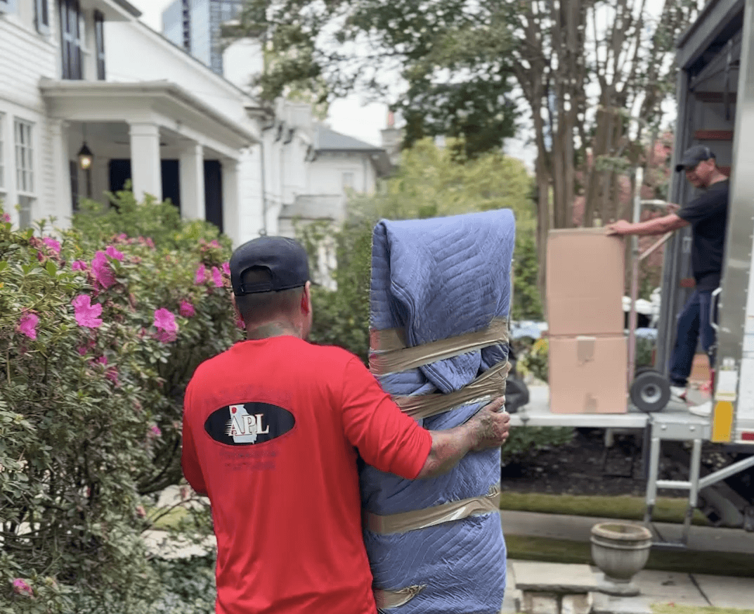 Mover in red shirt carrying a padded moving blanket, surrounded by greenery and flowers, with a moving truck in the background, illustrating Atlanta Pack and Load's reliable moving services.