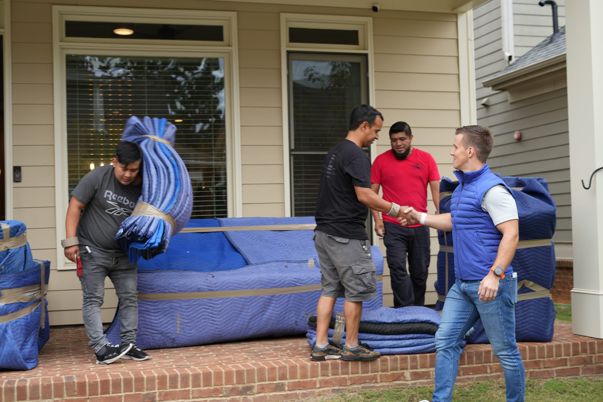 Movers in blue and black uniforms handling moving blankets, shaking hands in front of a house, showcasing Atlanta Pack and Load's commitment to reliable service and customer care.