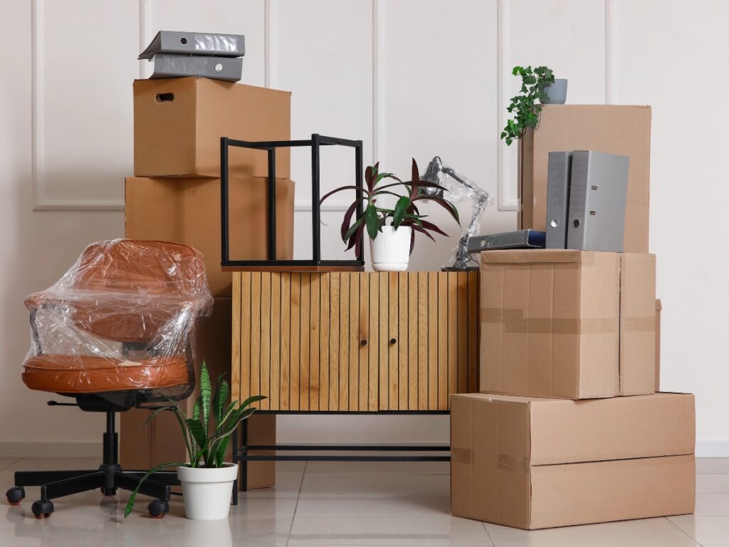 Moving supplies including stacked cardboard boxes, a wrapped office chair, and a wooden cabinet with a plant, representing essential elements for creating a moving budget and planning a relocation.