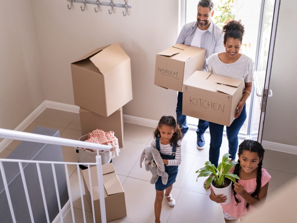 Family moving boxes labeled "Bedroom" and "Kitchen" into a new home, with children carrying a plant and stuffed animal, illustrating a DIY moving process.