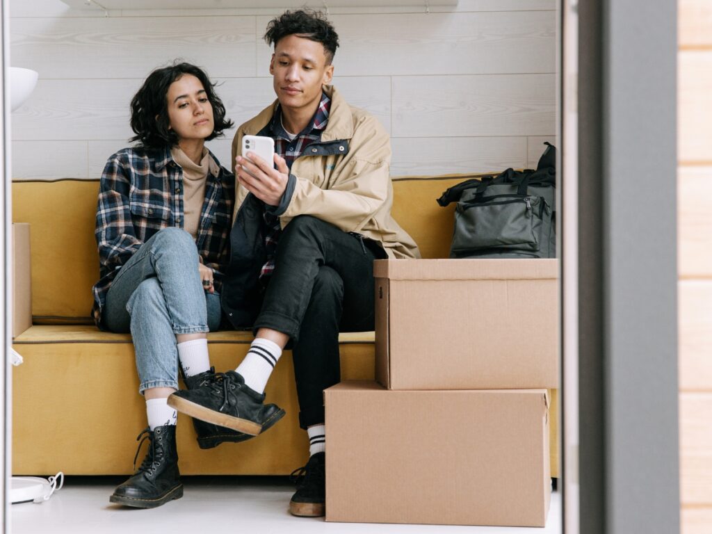 Couple sitting on a yellow couch surrounded by moving boxes, looking at a smartphone, reflecting the post-move budgeting and settling-in process.