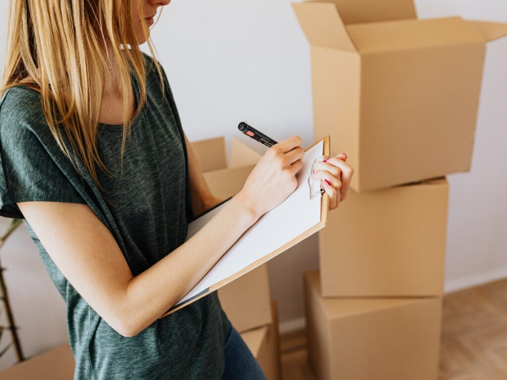 Woman writing on clipboard in front of stacked moving boxes, emphasizing organization and budgeting during relocation.
