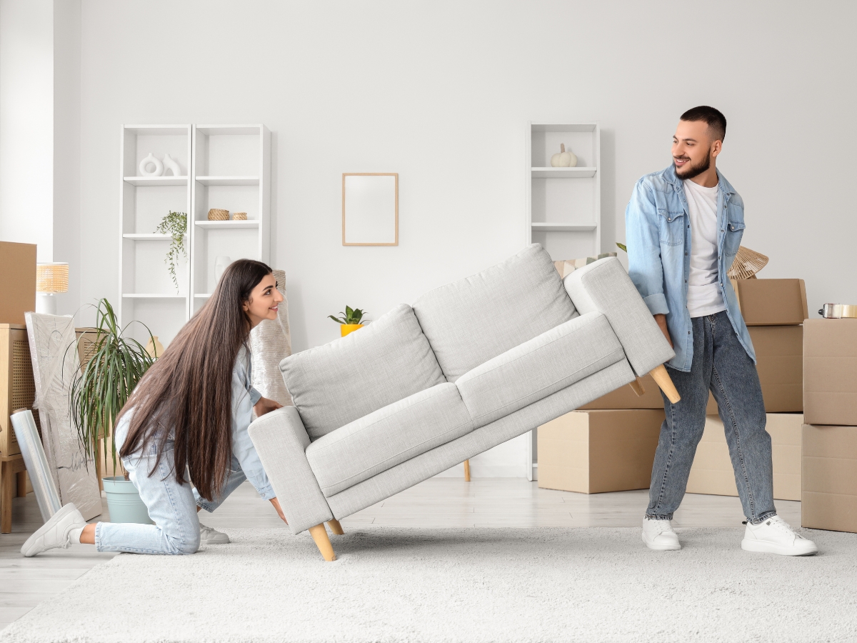 Couple moving a light gray sofa in a bright room filled with moving boxes, emphasizing teamwork in the relocation process.