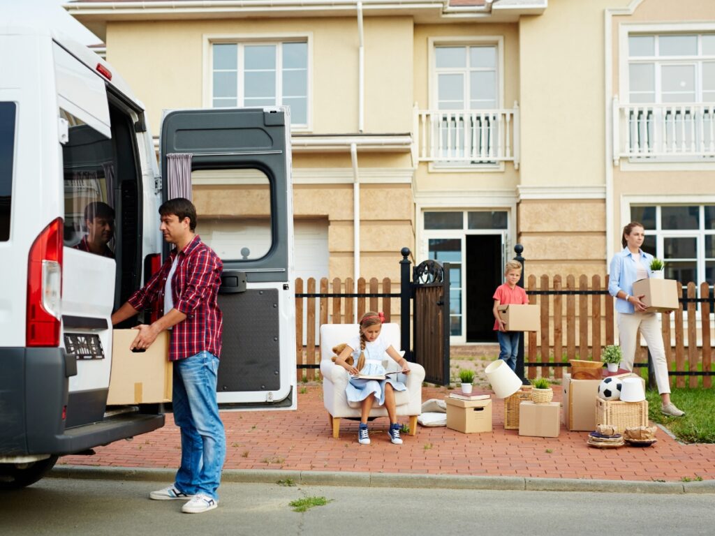 Family preparing for a long-distance move, loading boxes into a moving van, with a child sitting on a chair surrounded by packed items and plants, illustrating the logistics of relocation and the importance of moving insurance.