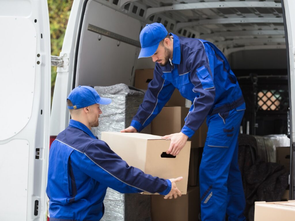 Two workers in blue uniforms use proper heavy lifting techniques as they load cardboard boxes into a van, with one man handing a box to another inside the vehicle.