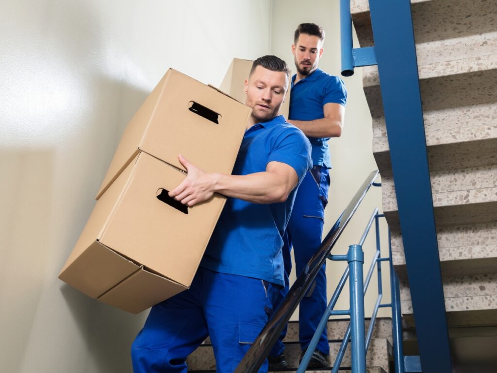 Two men in blue uniforms use proper heavy lifting techniques as they carry cardboard boxes down a stairwell.