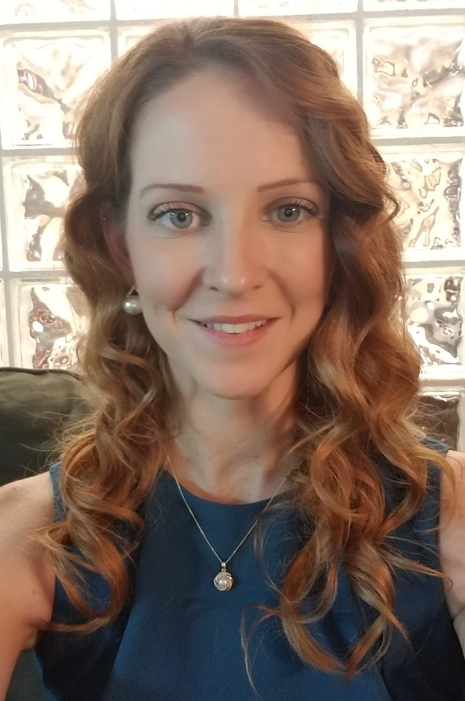 A woman with long, wavy auburn hair and blue eyes is smiling at the camera. She is wearing a blue sleeveless top, pearl earrings, and a pendant necklace&mdash;perfect for our About Us page.