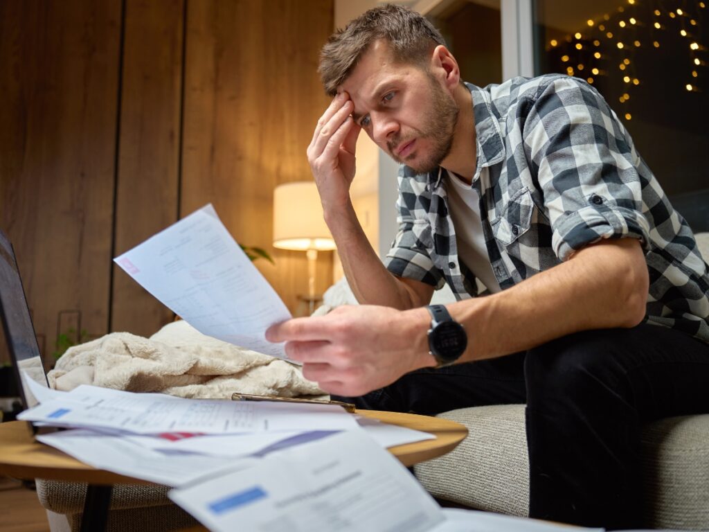 A man sits on a couch reviewing bills, looking concerned about potential utility disconnection, with paperwork spread out on the table in front of him.