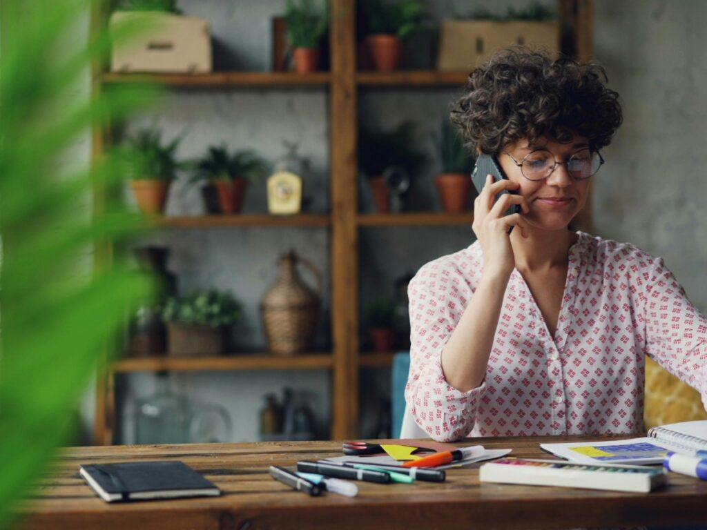 A person with curly hair and glasses sits at a wooden table, talking on the phone about a utility disconnection, surrounded by notebooks and pens, with plants on shelves in the background.