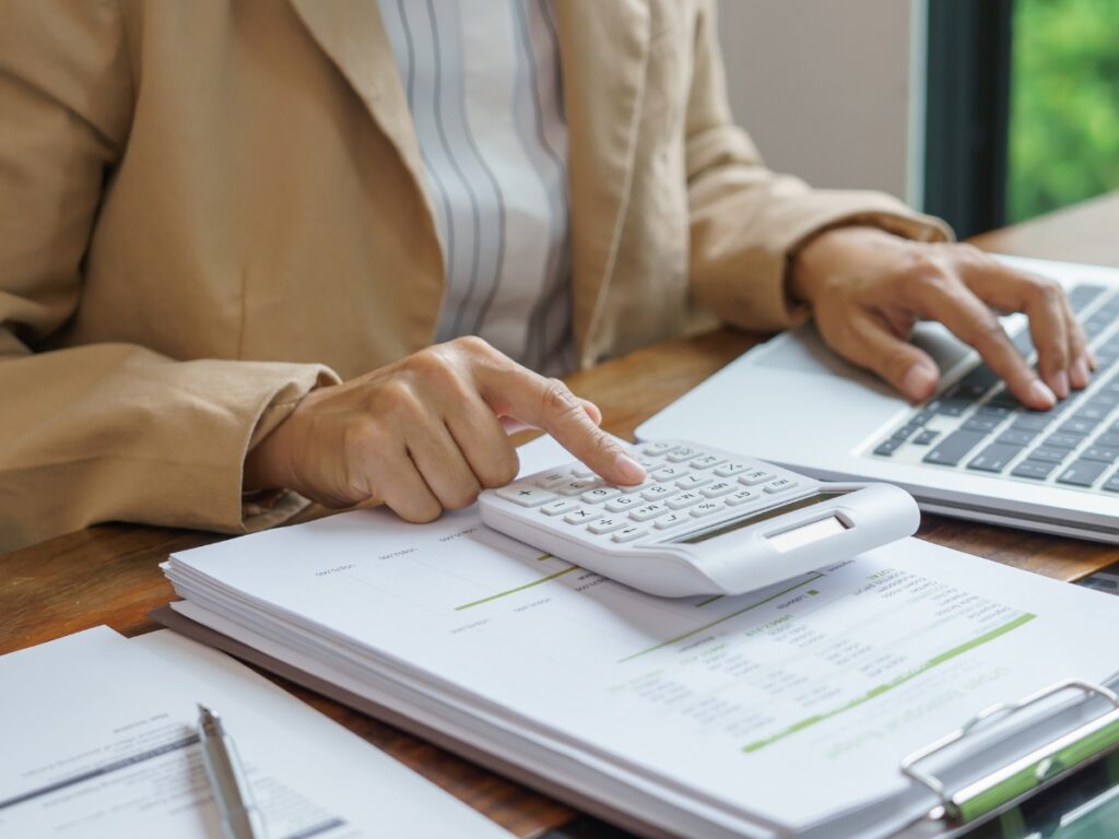 Person calculating expenses related to utility disconnection on top of financial documents while working on a laptop at a wooden desk.