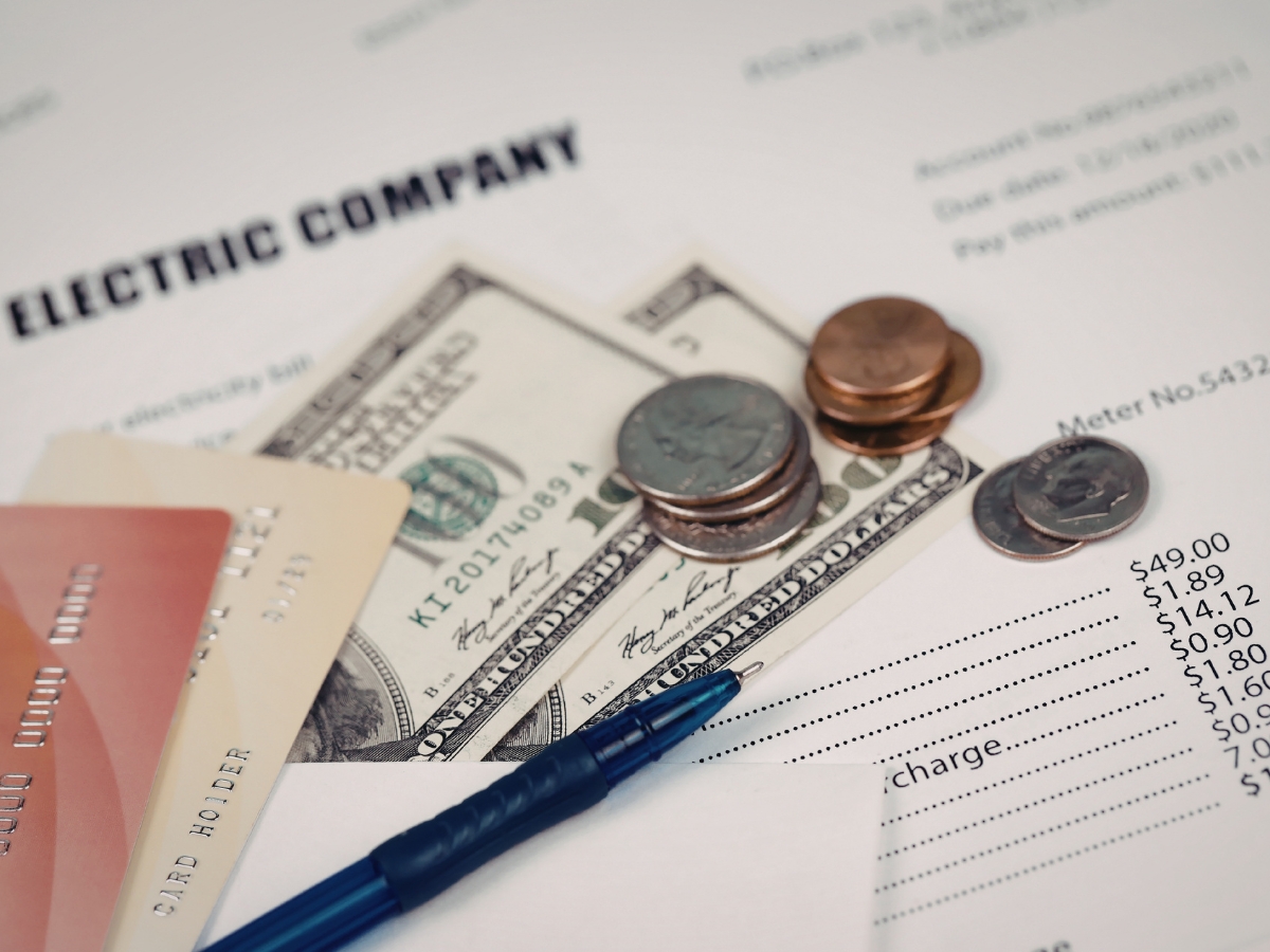 An electric bill with cash, coins, credit cards, and a pen placed on top, highlighting the urgency of payment to avoid utility disconnection.