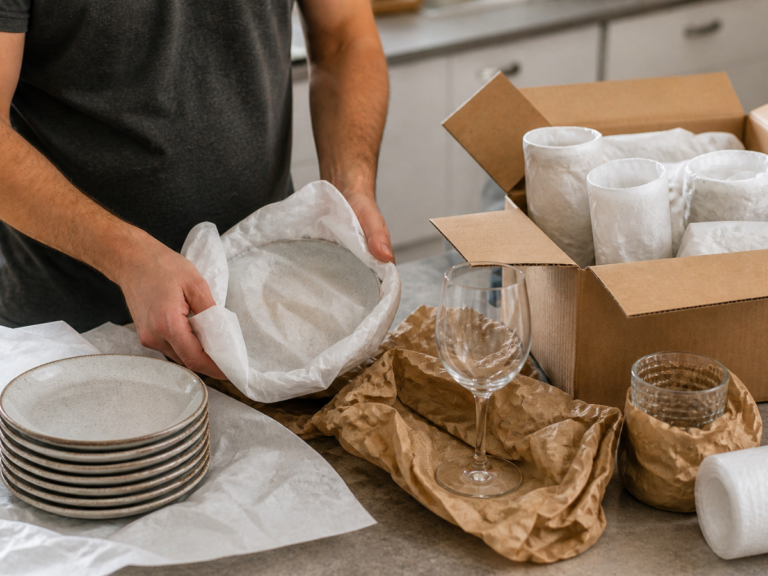 A person wraps fragile ceramic plates in packing paper next to a stack of plates, glassware, and a cardboard box on a kitchen counter, following essential packing tips for delicate items.
