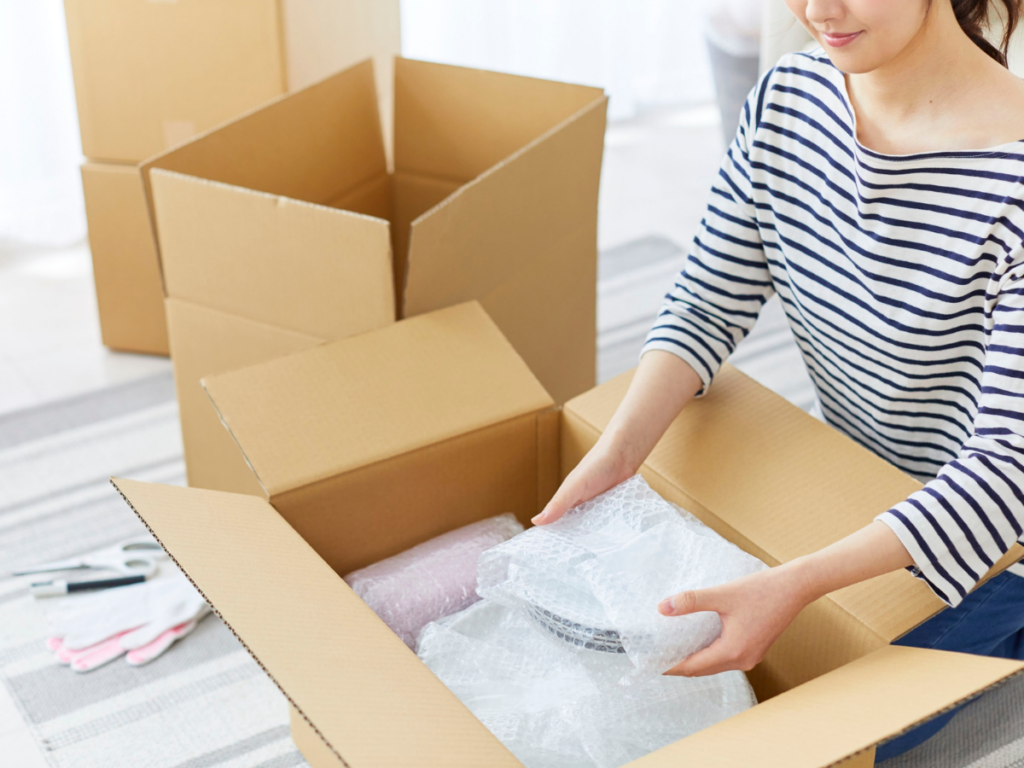 A person wearing a striped shirt packs fragile items into a cardboard box, following packing tips such as using bubble wrap, with other boxes and gloves visible nearby.