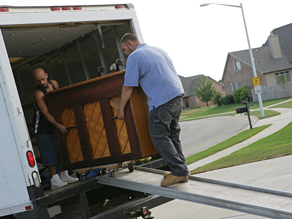 Two men load a wooden piano—a special items moving task—into a moving truck using a metal ramp on a suburban street.