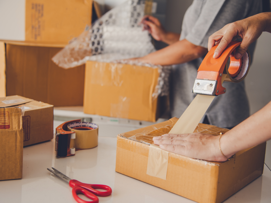 Two people packing cardboard boxes, one sealing a box with tape and the other using bubble wrap to protect special items for moving, with scissors and a tape dispenser on the table.