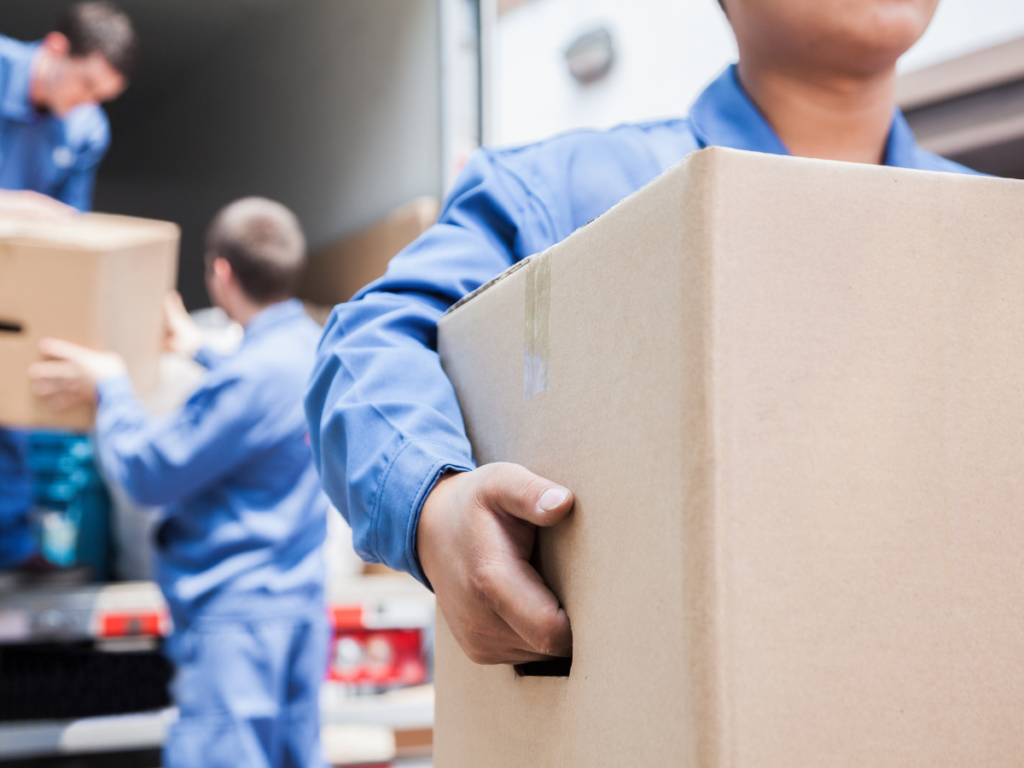 People in blue uniforms are carefully unloading special items from a vehicle, carrying cardboard boxes as part of a moving process.