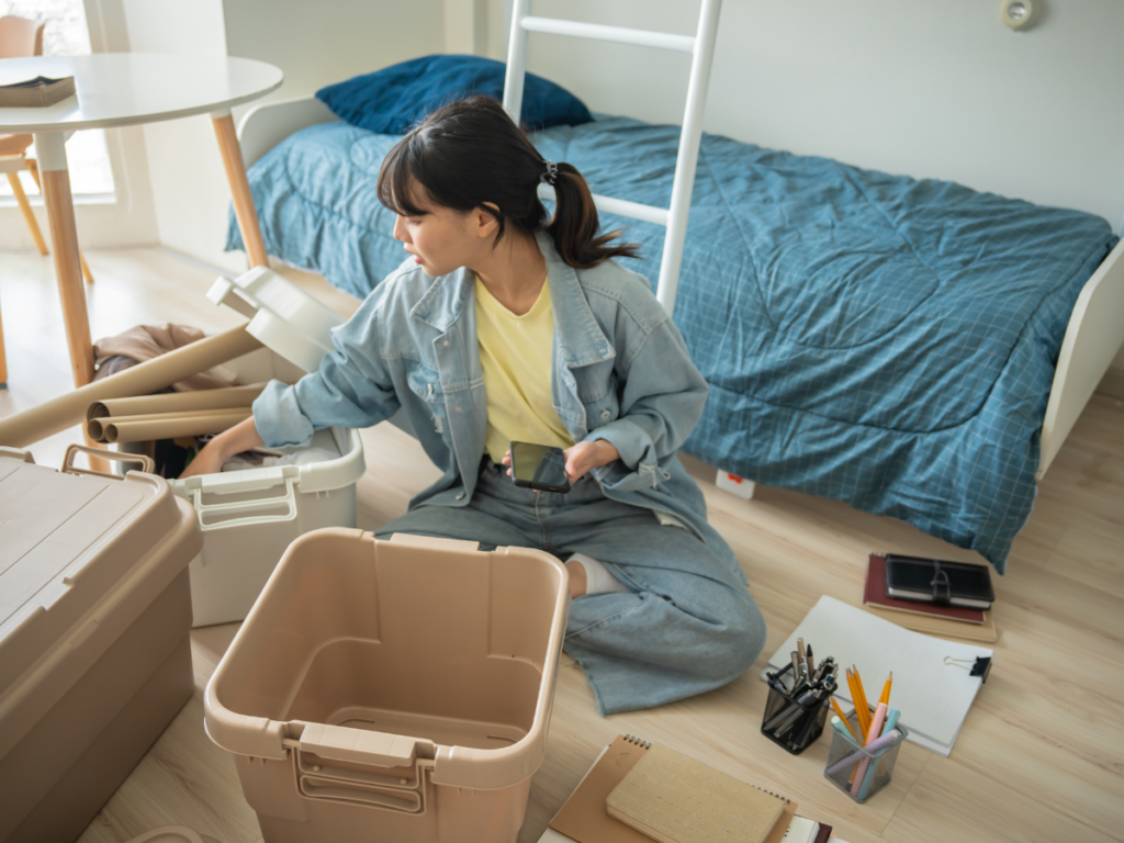 A person sits on the bedroom floor, using an unpacking strategy to sort items into plastic storage bins, with notebooks and stationery scattered nearby.
