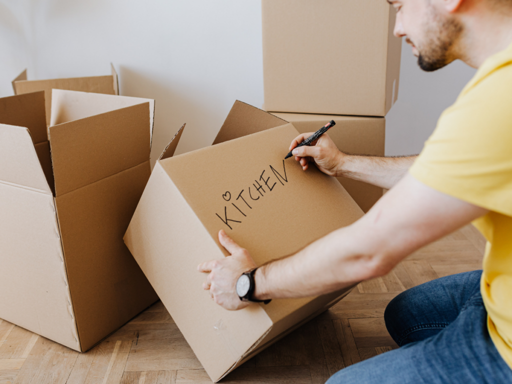 A person in a yellow shirt labels a cardboard box "KITCHEN" with a black marker, using an unpacking strategy while surrounded by other open moving boxes.
