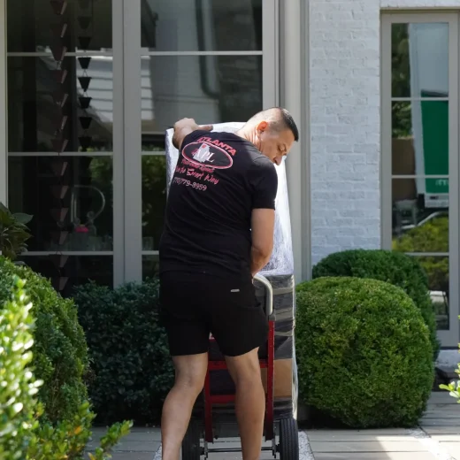 Mover in Atlanta Pack & Load shirt loading a dolly with wrapped furniture outside a home, surrounded by greenery, illustrating professional moving services.