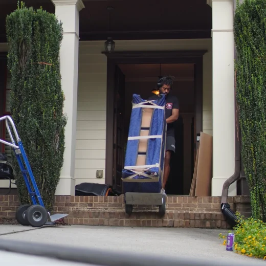 Man using a dolly to move a large, wrapped item from a porch, with packing materials and moving equipment visible, illustrating Atlanta Pack & Load's professional moving services.
