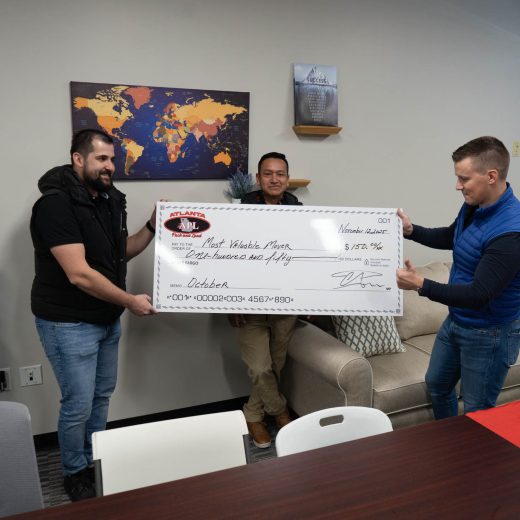 Three Atlanta Pack and Load team members celebrating with a large novelty check, recognizing the "Most Valuable Mover" award in an office setting, featuring a world map and modern decor.