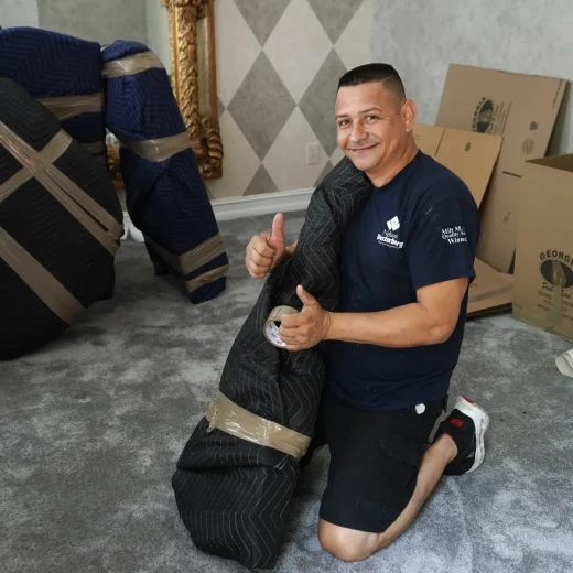 Man kneeling on carpet holding a rolled moving blanket, smiling and giving a thumbs-up, with moving boxes and padded furniture in the background, illustrating professional packing services for commercial moves.