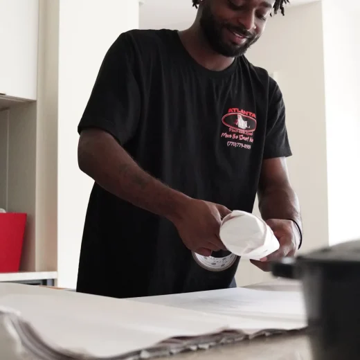 Man in a black Atlanta Pack and Load t-shirt carefully packing items with protective materials in a bright, organized workspace, illustrating professional packing and loading services for commercial moves.