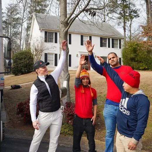 Group of four movers in casual attire, smiling and waving, outside a suburban home, emphasizing teamwork and a friendly service approach for packing and loading.