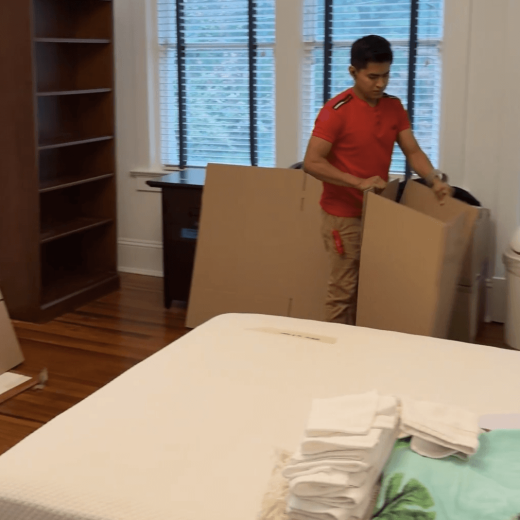 Person in red shirt packing boxes in a room, preparing for a move, with a bed and shelves visible, illustrating packing and loading services for relocation.